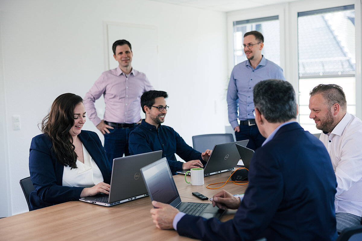 Besprechung in modernem Büro mit mehreren Mitarbeitenden an einem Konferenztisch, die gemeinsam an Laptops arbeiten und Ideen austauschen.